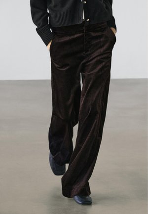 Person iført mørkebrune viddebenet kordfløjlsbukser, sorte sko og en sort, knappet sweater med hænderne i lommerne.
