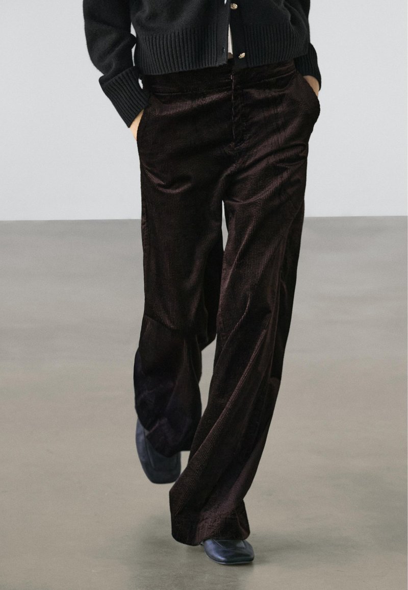 Person iført mørkebrune viddebenet kordfløjlsbukser, sorte sko og en sort, knappet sweater med hænderne i lommerne.