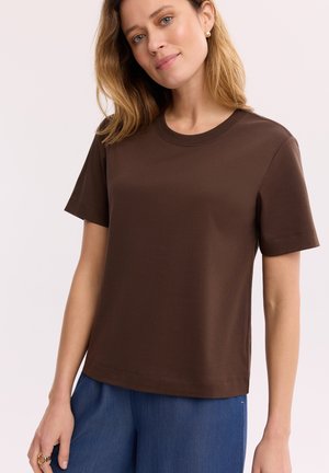 Młoda kobieta o blond włosach do ramion, ubrana w ciemnobrązową koszulkę z krótkim rękawem i niebieskie spodnie, stoi z lekkim uśmiechem.