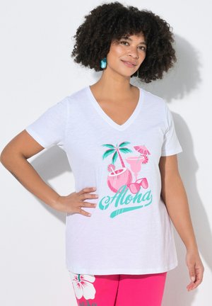 Frau mit lockigem Haar, die türkisfarbene Ohrringe trägt, ein weißes V-Ausschnitt-Shirt mit pink-grünem Tropenmuster und dem Schriftzug "Aloha" sowie eine pinke Hose.