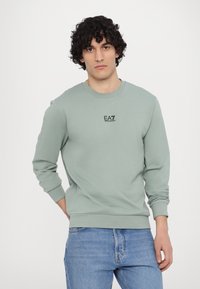 Felpa verde menta realizzata in tessuto morbido, con collo rotondo, maniche lunghe e un piccolo logo EA7 nero sul petto. Indossata con jeans blu.
