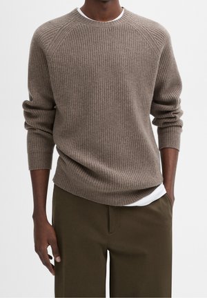 Mand iført en brun ribstrikket sweater over en hvid skjorte, kombineret med mørke olivengrønne bukser, stående med den ene hånd i lommen.