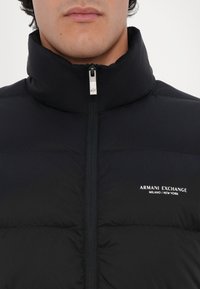 Sort pufferjakke med høj krave, udstyret med lynlås foran og "Armani Exchange" logo på brystet. Glat tekstur, let design.