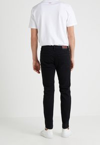 Man i svarta slim-fit jeans, vita sneakers och en vit T-shirt med "HUGO"-logotyp på baksidan av kragen och midjan, som står mot en vit bakgrund.