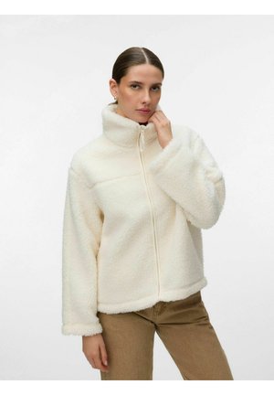Femme portant une veste zippée duveteuse de couleur crème avec un col montant et un pantalon marron sur un fond blanc uni.
