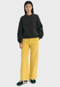 Giovane donna in piedi che indossa un maglione a coste marrone scuro, pantaloni a gamba larga in corduroy giallo e sneakers nere con lacci bianchi.
