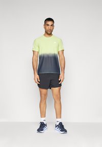 Camiseta deportiva de manga corta con un degradado de verde lima a azul marino oscuro, combinada con pantalones cortos negros y zapatillas de running en azul marino con detalles en blanco.