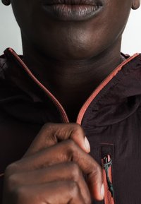 Close-up van het onderste deel van iemands gezicht en een hand die de rits van een donker jasje met koraalkleurige rand omhoog trekt.