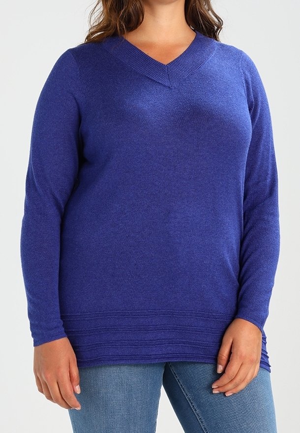 Femme portant un pull bleu à manches longues et col en V, associé à un jean bleu, debout les mains détendues le long du corps.