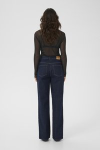 Vrouw met golvend bruin haar, gekleed in een doorzichtig zwart T-shirt met lange mouwen en hoge taille donkerblauwe wijde jeans, staande met haar rug naar de camera.