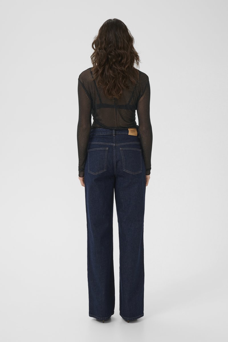 Frau mit welligem braunem Haar, die ein transparentes schwarzes Langarm-Oberteil und hochgeschnittene dunkelblaue, weit geschnittene Jeans trägt, steht mit dem Rücken zur Kamera.