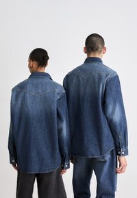 Denimskjorter i mørk blå med en falmet tekstur. Har en spiss krage, lange ermer og knappefester. Bakdetaljene inkluderer skjorteveske-design.