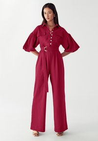 Burgundijos spalvos šokinėjanti suknelė su pūstomis rankovėmis, mygtukų užsegimu, dviem krūtinės kišenėmis ir juosmeniu su diržu. Lygi medžiaga su plačiais džinsais.