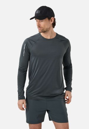 Mand iført en sort kasket, mørkegrå langærmet skjorte og matchende mørkegrå atletikshorts, kigger mod venstre.