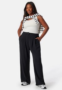 Pantalons larges noirs à motif plissé, associés à un débardeur blanc ajusté et à un pull en maille rayé porté sur les épaules.