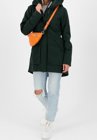 Tmavě zelená nepromokavá bunda s kapucí, která má dvě přední kapsy. Nahoře má zlight blue roztrhané džíny a je doplněna oranžovou přes rameno taškou.