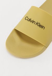 Sandală din cauciuc galbenă, cu talpă plată, care prezintă o curea lată cu suprafață texturată și un logo „Calvin Klein” embosat în negru. Design cu vârful rotunjit.