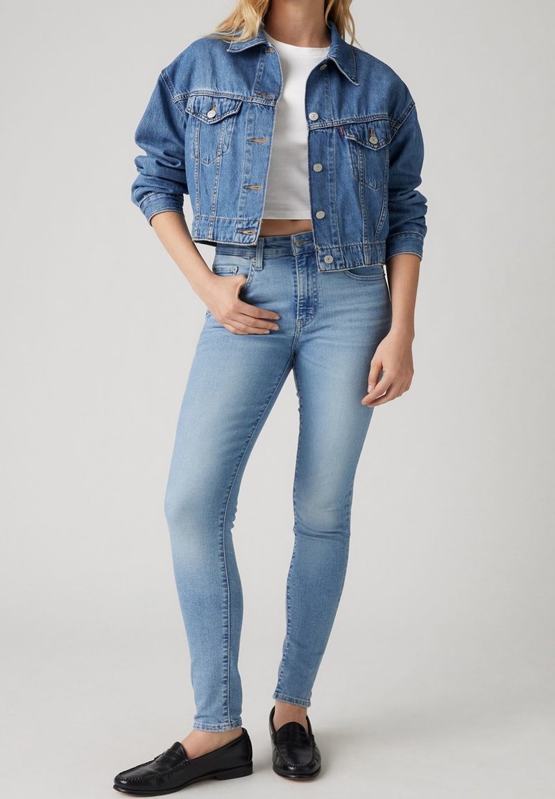 Femme portant une veste en jean courte, un crop top blanc, un jean skinny bleu clair et des mocassins en cuir noir, debout devant un fond uni.