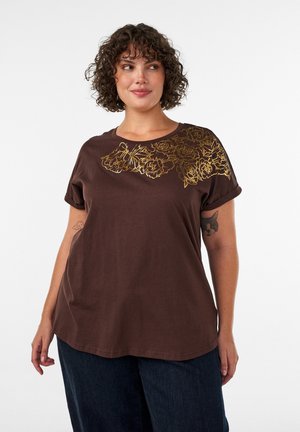Γυναίκα με σγουρά μαλλιά, που φορά ένα καφέ t-shirt με χρυσό floral σχέδιο στον ώμο και σκούρα μπλε τζιν, στέκεται μπροστά σε λευκό φόντο.