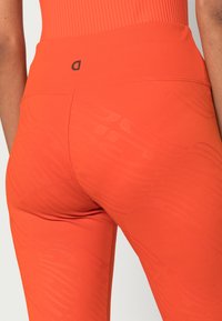 Leggings atléticos laranja apresentam uma cintura alta, textura lisa e um padrão tonal subtil ao longo do tecido. Logotipo preto na cintura.