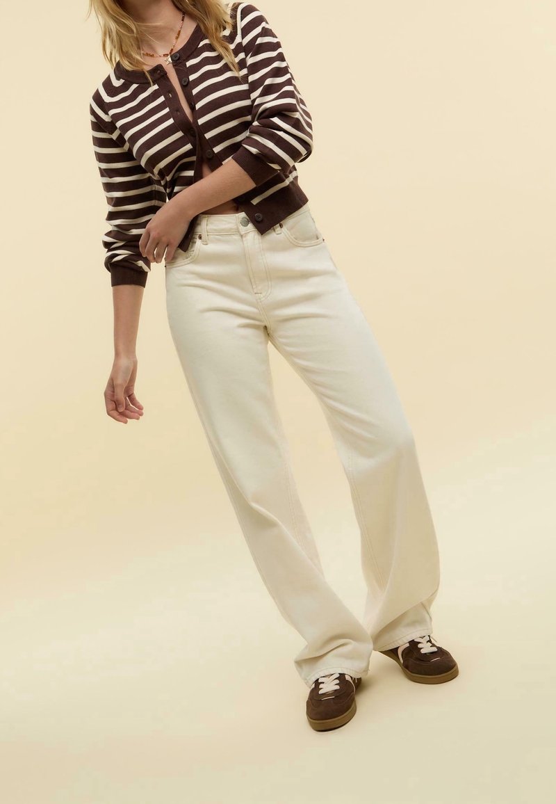 Femme portant un cardigan rayé marron et blanc, un pantalon large crème, et des chaussures marron à lacets sur un fond beige clair.