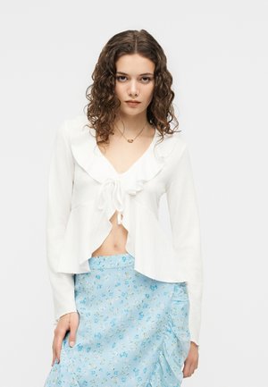 Žena s kučeravými hnedými vlasmi má na sebe biely nariasený top s dlhými rukávmi zviazaný vpredu a svetlomodrú kvetovanú nariasenú sukňu, pózujúc na jednoliatom pozadí.