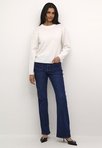Hvid strikket sweater med Rund hals, parret med mørkeblå bredbundede jeans. Modellen har sorte spidse hæle på. Ensfarvet baggrund.