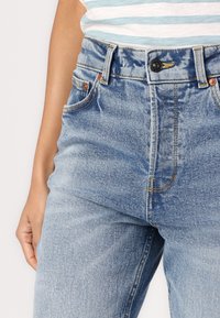 Ljusblå högmidjade jeans med en blekt, texturerad yta. Har knappstängning, kopparnitar och sidofickor.
