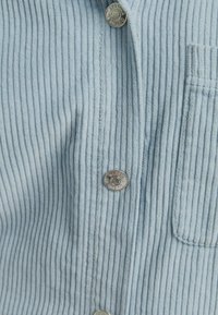 Camicia in velluto a coste blu chiaro con textura a righe verticali, bottoni a pressione argentati e taschino sul petto.