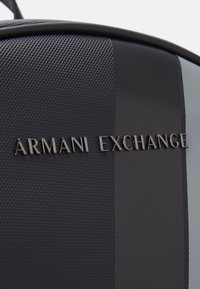 Zaino nero in tessuto con sezioni lisce e decorate. Il nome del marchio "ARMANI EXCHANGE" è impresso in elegante hardware nero.