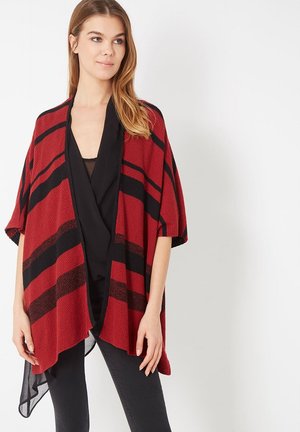 Mujer que lleva un chal suelto de rayas rojas y negras sobre una blusa negra y pantalones negros, de pie frente a un fondo blanco liso.