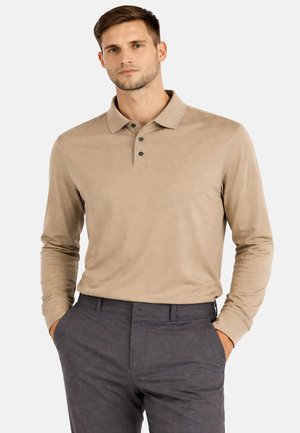 Man draagt een beige poloshirt met lange mouwen en donkere knopen, samen met grijze broek, staand met handen in de zakken tegen een effen achtergrond.