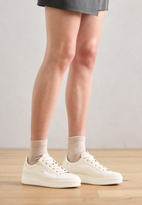 Hvite lær sneakers med gullfargede detaljer, avrundet tå og snøring. Kombinert med ribbestrikkede beige sokker, satt mot et tregulv og grå bakgrunn.