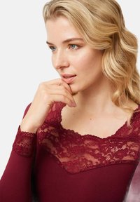 Top burdeos con escote de encaje y mangas largas. El encaje presenta patrones florales, tejido texturizado y un corte en forma de V en el cuello.