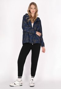 Marinblå och mörkgrön kamouflagejacka med huva, dragkedjestängning och elastiska ärmslut, kombinerad med svarta joggers och vita sneakers.