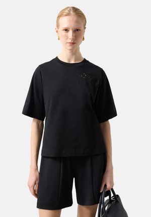 Kvinde i sort oversized T-shirt og knælange shorts holder sort håndtaske mod en ensfarvet lys baggrund.