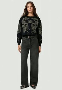 Mulher a usar um suéter preto com padrões de serpente e flores, jeans escuros de cintura alta, cinto preto e sapatos pretos de bico.
