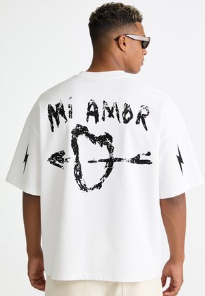Uomo che indossa una maglietta bianca oversize con testo nero "Mi Amor" e grafica di un cuore trafitto da una freccia sulla schiena e fulmini sulle maniche.