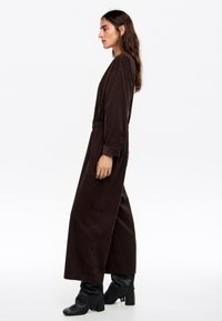 Mono marrón de manga larga con un corte relajado, cintura elástica, bolsillos laterales, tejido texturizado y piernas anchas, combinado con botas de tacón negras.