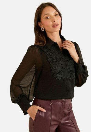 Blusa nera trasparente con dettagli in pizzo ricamato sul davanti, maniche a sbuffo e chiusura a bottoni, abbinata a pantaloni in ecopelle bordeaux.