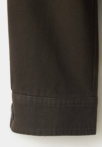 Jambe de pantalon en tissu brun foncé avec des coutures visibles et un petit bouton près du poignet sur un fond clair.
