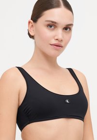 Black bralette made from smooth fabric, featuring a rounded neckline, wide straps, and a small CK logo at the center.