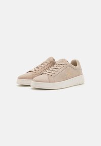 Adidași din suede bej cu tălpi din cauciuc alb. Caracteristicile includ șireturi plate, vârf rotunjit și un accent cu logo pe lateral.