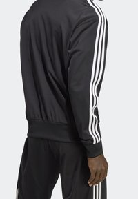 Svart idrottsjacka och byxor med vita trippla ränder på ärmarna och benen, bärs av en stående person med mörk hud.