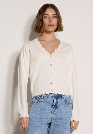 Donna che indossa un cardigan bianco con bordi smerlati e bottoni, maniche lunghe e jeans azzurri a vita alta, in piedi davanti a uno sfondo semplice.