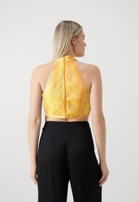 Gelbes Halter-Crop-Top mit glatter Textur, ausgestattet mit einem Reißverschluss auf der Rückseite. Kombiniert mit schwarzen weit geschnittenen Hosen mit vertikalen Streifen.