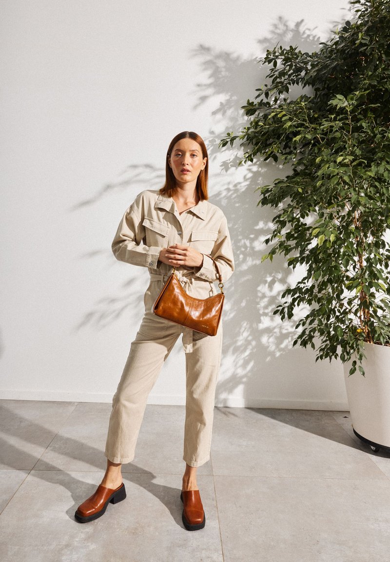 Tenue beige style cargo à manches longues, sac en cuir marron et sabots à talons marron. Mur blanc et verdure en arrière-plan.
