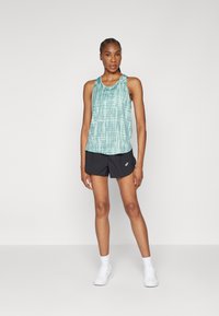 Kvinne står iført en tanktop med teal mønster, svarte treningsshorts, hvite sokker og hvite joggesko mot en ensfarget lys bakgrunn.
