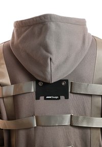 Chaqueta gris con capucha que presenta un tejido texturizado, correas ajustables beige y una etiqueta de cuero negro con "MWM Design" en texto en relieve.