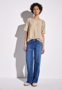 Beige tričko s krátkým rukávem s hladkou texturou, kombinované s volnými modrými džíny. Modelka stojí na jednobarevném bílém pozadí.